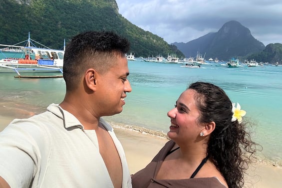 Trisha & Nikhil standing on El Nido Beach. Looking at each other with clear sea, mountains and boats