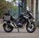 A custom motorcycle with a rugged appearance is parked on a dirt path. The bike is designed with a matte black paint job, featuring a branded logo of 'Cafe Racer'. It is equipped with dual front suspensions, a circular headlamp with grill protection, and handlebars extending outward. The background includes residential buildings and some trees.