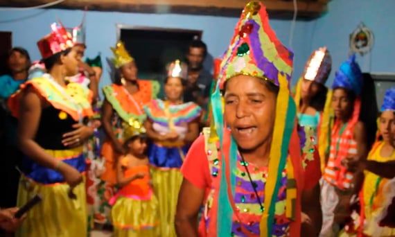 Mulheres e crianças com trajes e coroas coloridas cantam e dançam o Reizado em uma casa decorada