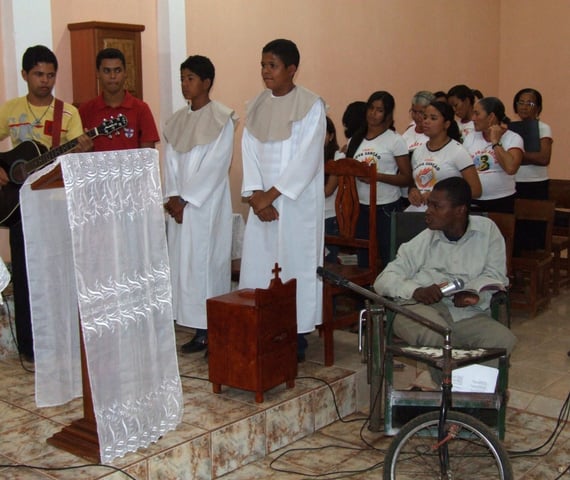 Pessoas celebram São João na igreja, com trajes religiosos e fiel em cadeira de rodas