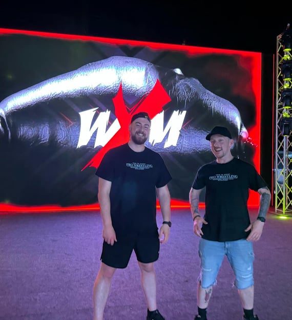 Two men stand in front of an LED screen at a wrestling show.