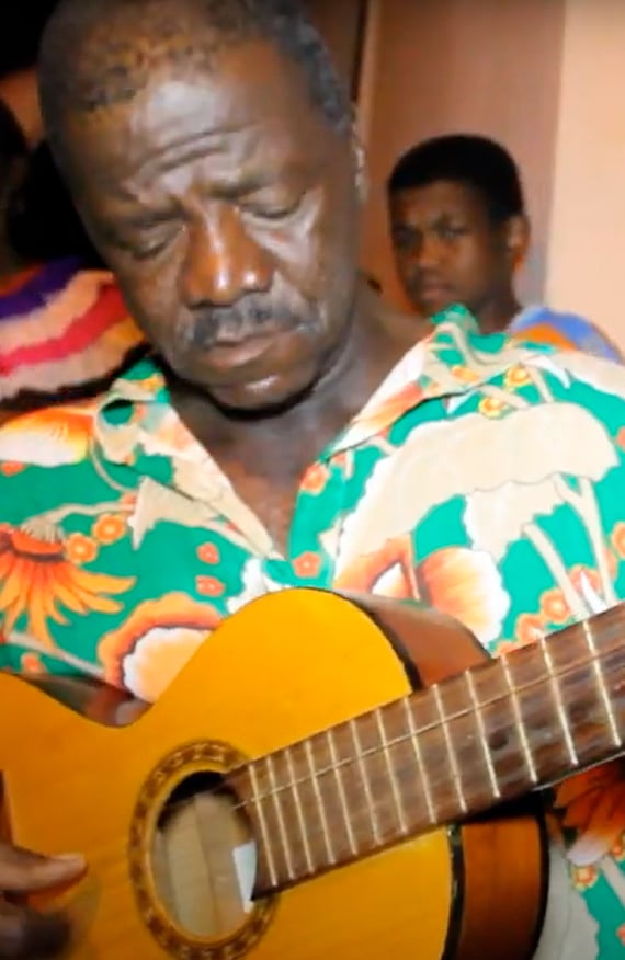 Sr. João Reis toca violão durante o Reizado, vestindo camisa florida, símbolo da tradição quilombola