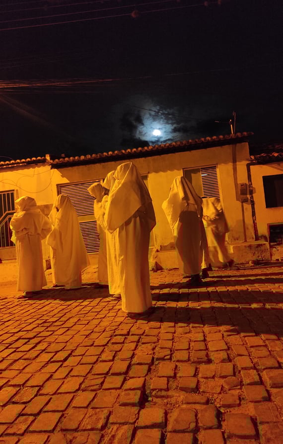 Penitentes cobertos de branco caminham à noite sob a luz da lua em rua de paralelepípedo