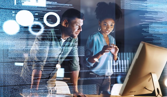 A male and female leader look at a computer monitor as holographic coding and scripts float around them.