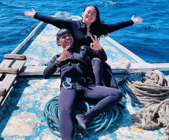 Sugbu Diver in a wetsuit with full scuba gear lying at the tip of the boat, raising her hands wide