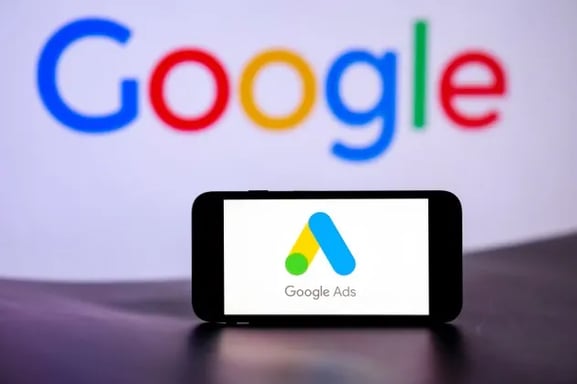 Celular en primer plano mostrando Google Ads, con una computadora desenfocada al fondo con "Google"