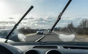 View from inside a car as windshield wipers clear washer fluid while driving on a sunny day with light traffic ahead.