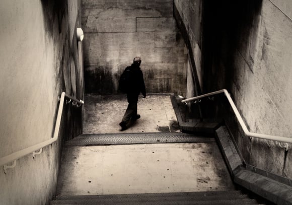 Photograph of a lone man dressed in black walking down a dimly lit subway staircase in London, captured in the early morning 