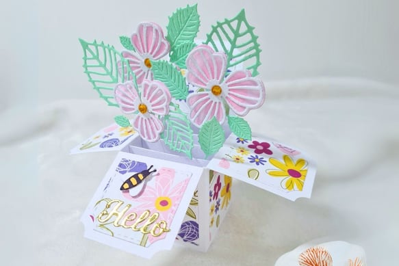 Pop-up card shaped like a box with pastel pink flowers and green leaves. "Hello" in gold script on a