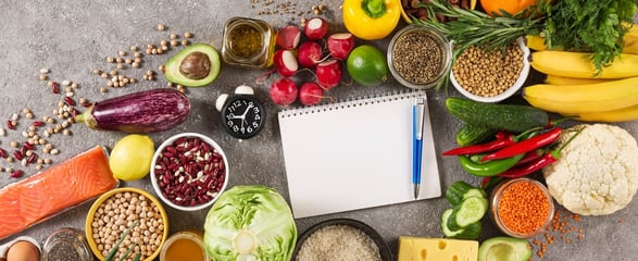A colourful array of healthy food, vegetables and grains, with a notepad and pen and a clock in the middle