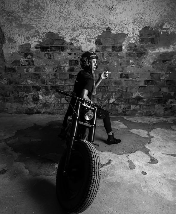 Modelo con casco y camiseta negra, posando sobre motocicleta clásica en espacio industrial