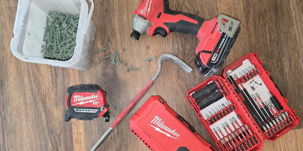 Milwaukee tools laid out on a hardwood floor with a box of nails