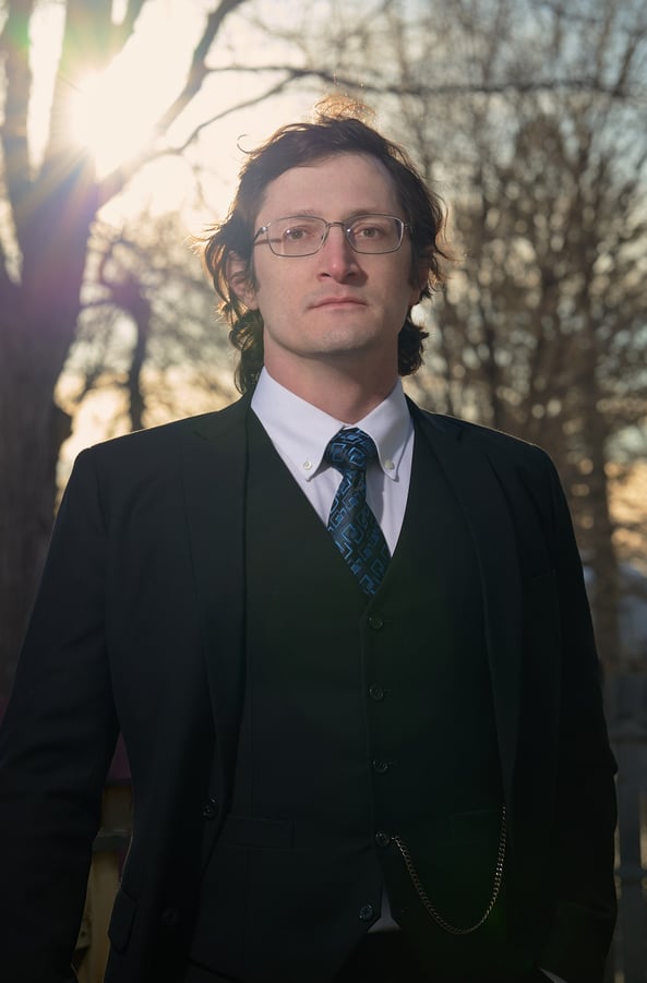 a man in a  black suit and glasses and tie with a blue tie in front of the sun and trees