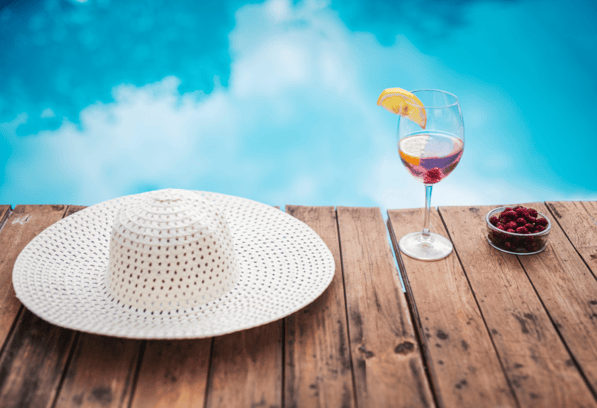 Un verre de rosé avec tranche de citron et un chapeau de plage blanc posés au bord d'une piscine 