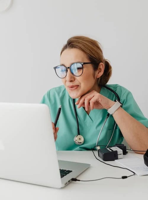 a woman in a green scrubsuit and glasses is looking at a laptop