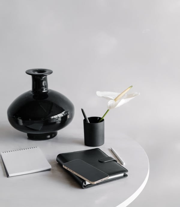 A black vase, pencil cup with a a pen and flower in it, and notebooks on a table