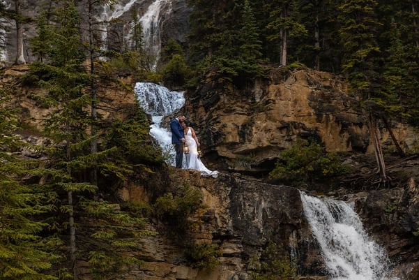 Bride and groom standing on a cliff next to a waterfall in Banff