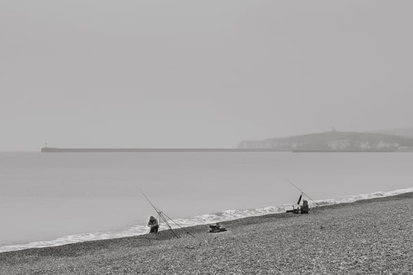 A black & white photo of two fishermen fishing on the back in seaford. Newhavens long stone pier and light house visible