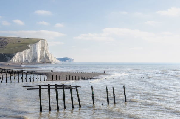 People walk along the beach in sussex between the old broken groynes with the chalk cliffs stretching into the distance