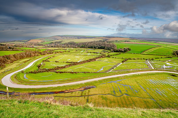 The Cuckmere river snakes through flooded green fields, hills in the background are shaded by dark cloud gathering