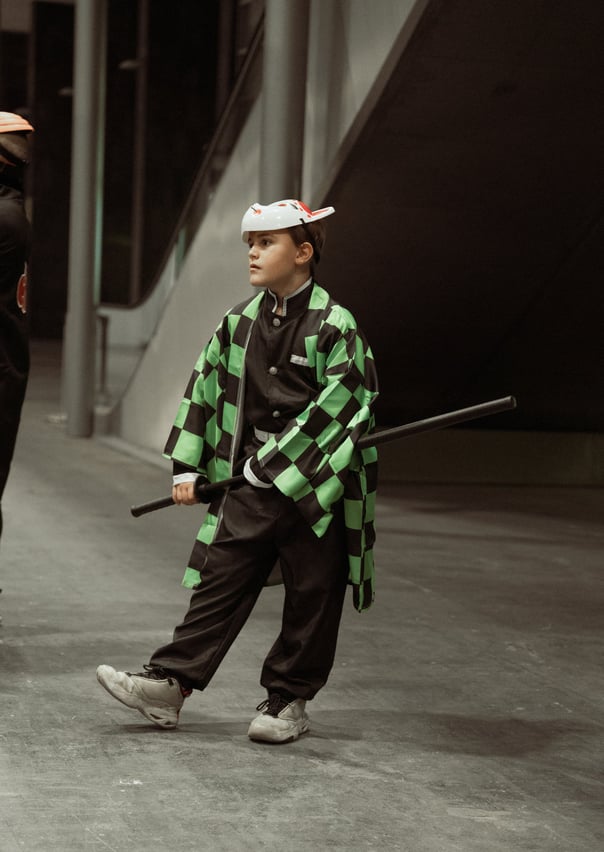 Young boy cosplaying Tanjiro Kamado from Demon Slayer, wearing a green and black checkered haori and a fox mask, holding a sw