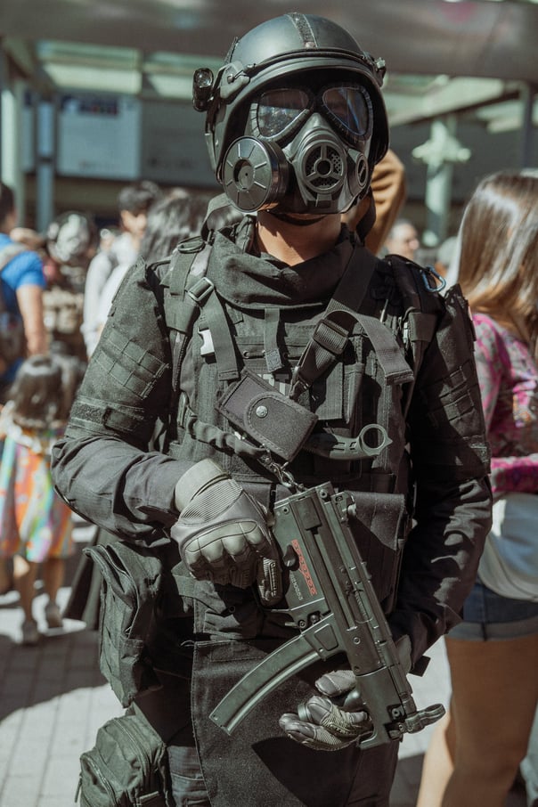 Cosplayer in full tactical black gear, including helmet and gas mask, holding a prop gun at IFEMA Madrid. Convention cosplay