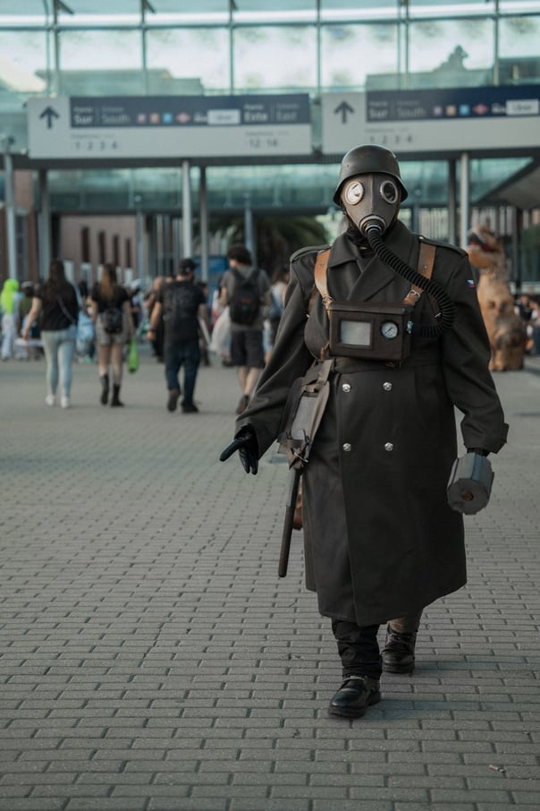 Cosplayer in a dark, military-style outfit with a gas mask and helmet walking on a paved surface. Convention cosplay