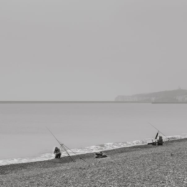 A black & white photo of two fishermen fishing on the back in seaford. Newhavens long stone pier and light house visible