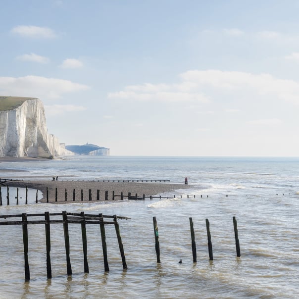 People walk along the beach in sussex between the old broken groynes with the chalk cliffs stretching into the distance