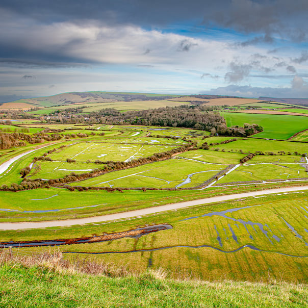 The Cuckmere river snakes through flooded green fields, hills in the background are shaded by dark cloud gathering