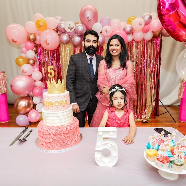 A young girl named Aurora celebrates her fifth birthday wearing a pink dress, standing between her parents