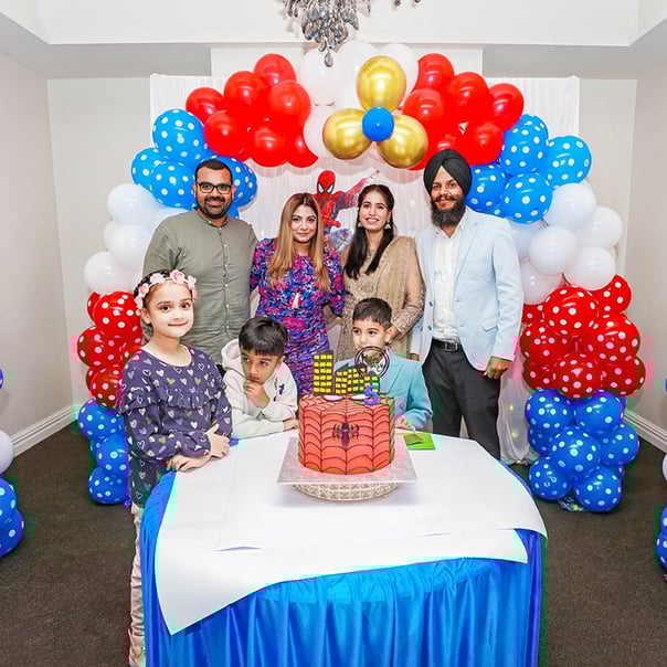 A birthday celebrant surrounded by a group of people, standing behind a colorful cake with vibrant decorations in the back.