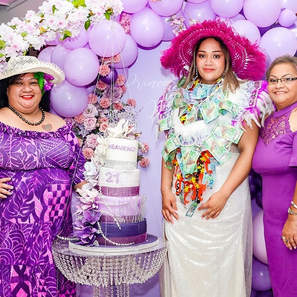 Three young women stand with a beautiful birthday cake, set against a purple background, creating a festive atmosphere.