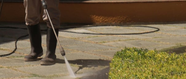 person in wellies uses pressure washing wand to clean stone floor near an hedge