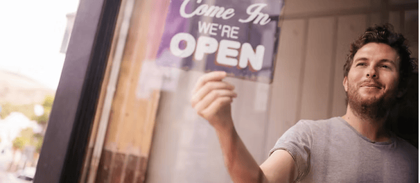 Man turning around Come In We're Open sign on coffee shop window