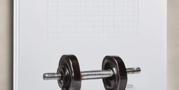 A kitchen countertop features various healthy food items including protein bars on a slate tray, a jar of yellow hummus, a wooden spoon, and a mug with a heart design. Packages of Alver Easy Protein powder are visible in the background. Fresh herbs, a bowl of blueberries, a container with seeds, strawberries, cherry tomatoes, and an avocado are also present, suggesting a focus on natural ingredients.