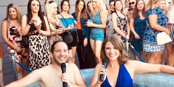 Alex Duffy and Ellie Brennan are in a hot tub with microphones, with audience behind.