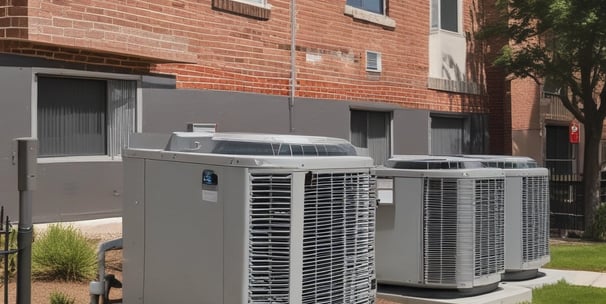 A view of a residential building with numerous air conditioning units attached to the exterior walls. The building features a pattern of square tiles and several balconies with various items placed outside. Palm trees and other greenery are visible in the foreground, indicating an urban environment with landscaped areas. Part of the image is framed by vertical bars and a chain.