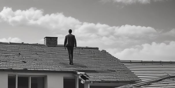 A person in a bright orange uniform and yellow hard hat is power washing the exterior of a structure next to a sidewalk. Large trees with dense green foliage are visible, along with bicycles parked under a canopy. The scene appears to be in an urban setting with buildings and construction in the background.