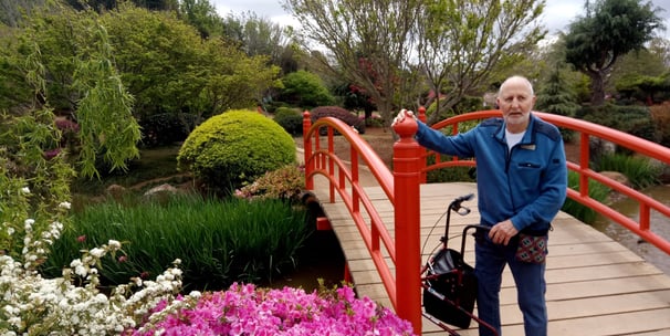 Older man standing at the foot of a beautiful red bridge in an ornamental garden with his walker