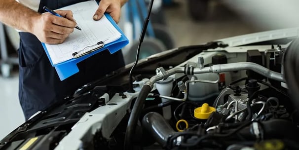 man with blue clipboard evaluating a car's engine.