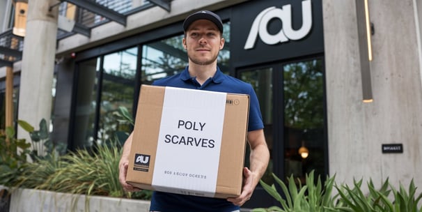 A photo of a man delivering boxes to an Au customer. The box is labeled Poly Scarves