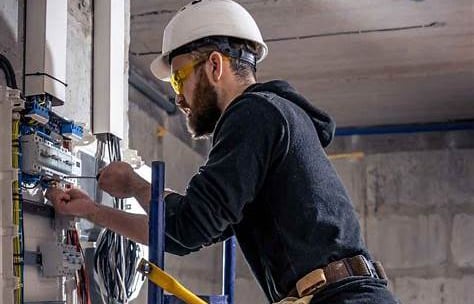 Alt="Commercial electrician installing wiring in a business facility, wearing hard hat and safety."