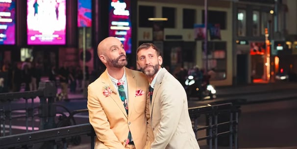 Pareja gay feliz tocándose la frente y sonriendo en una calle metropolitana de España, luz natural y momento espontáneo