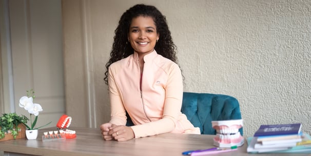 Mujer joven con cabello rizado sentada en un escritorio, sonriendo y rodeada de libros, modelos dentales y plantas en una ofi