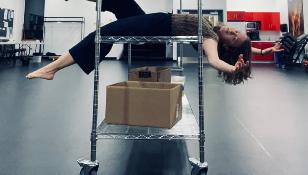 A young woman balances on a metal shelving unit, extending her limbs out and looking at the camera