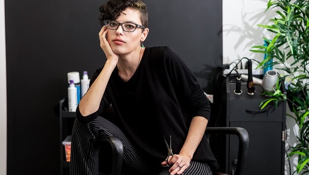A hairdresser sits in a salon chair holding posing with a pair of scissors