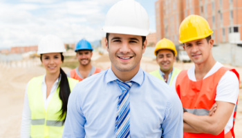 Engenheiro sorridente usando capacete branco e terno, com uma equipe de trabalhadores da construção civil ao fundo em um cant