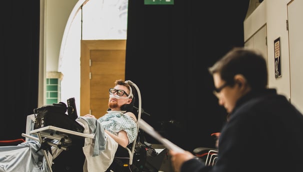 An electric wheelchair user with ventilator mask and a white BSL signer deep in conversation