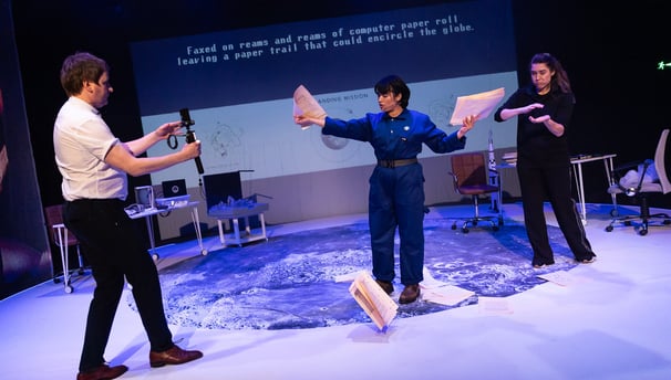 A white man, an Iranian woman in blue overalls and a BSL interpreter throw paper on the moon